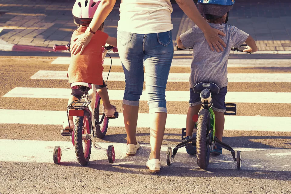 Close-up from back of woman walking on crosswalk with two small children on either side of her riding bicycles with training wheels.
